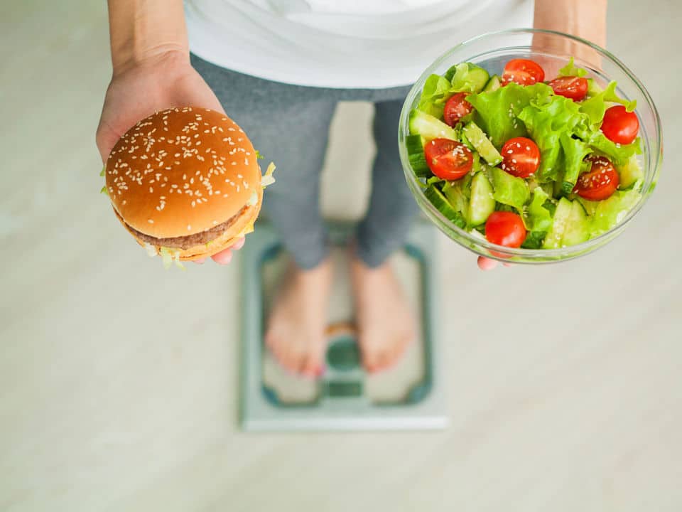woman on weighing scale holding burger and vegetable salad